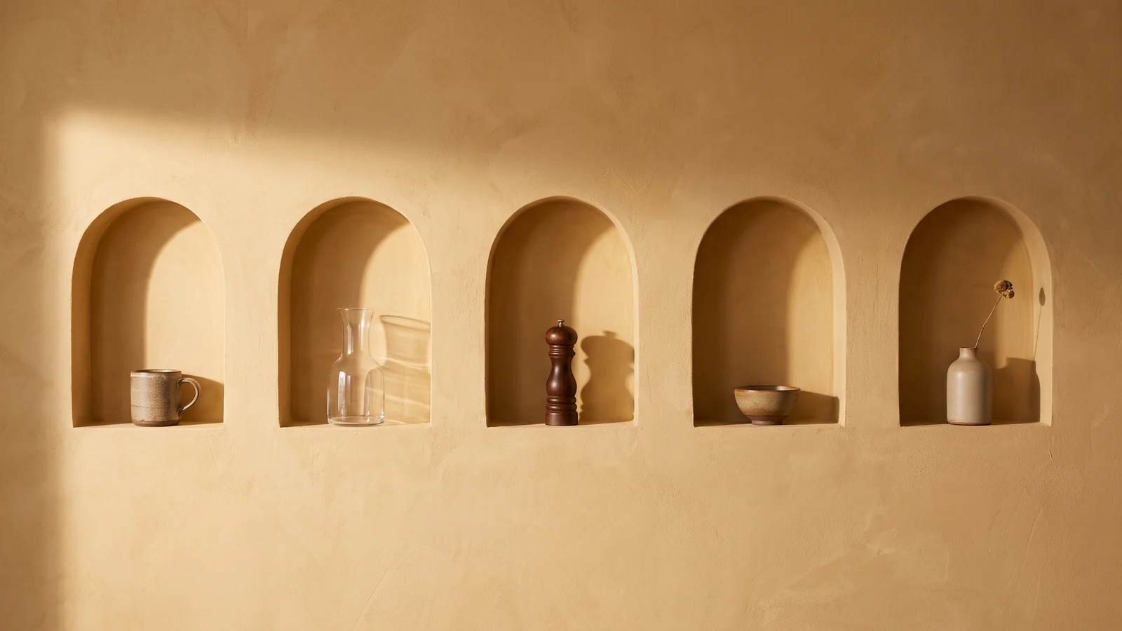 A warm plaster wall with a row of identical arched niches, each lit the same way and holding a different kitchen object